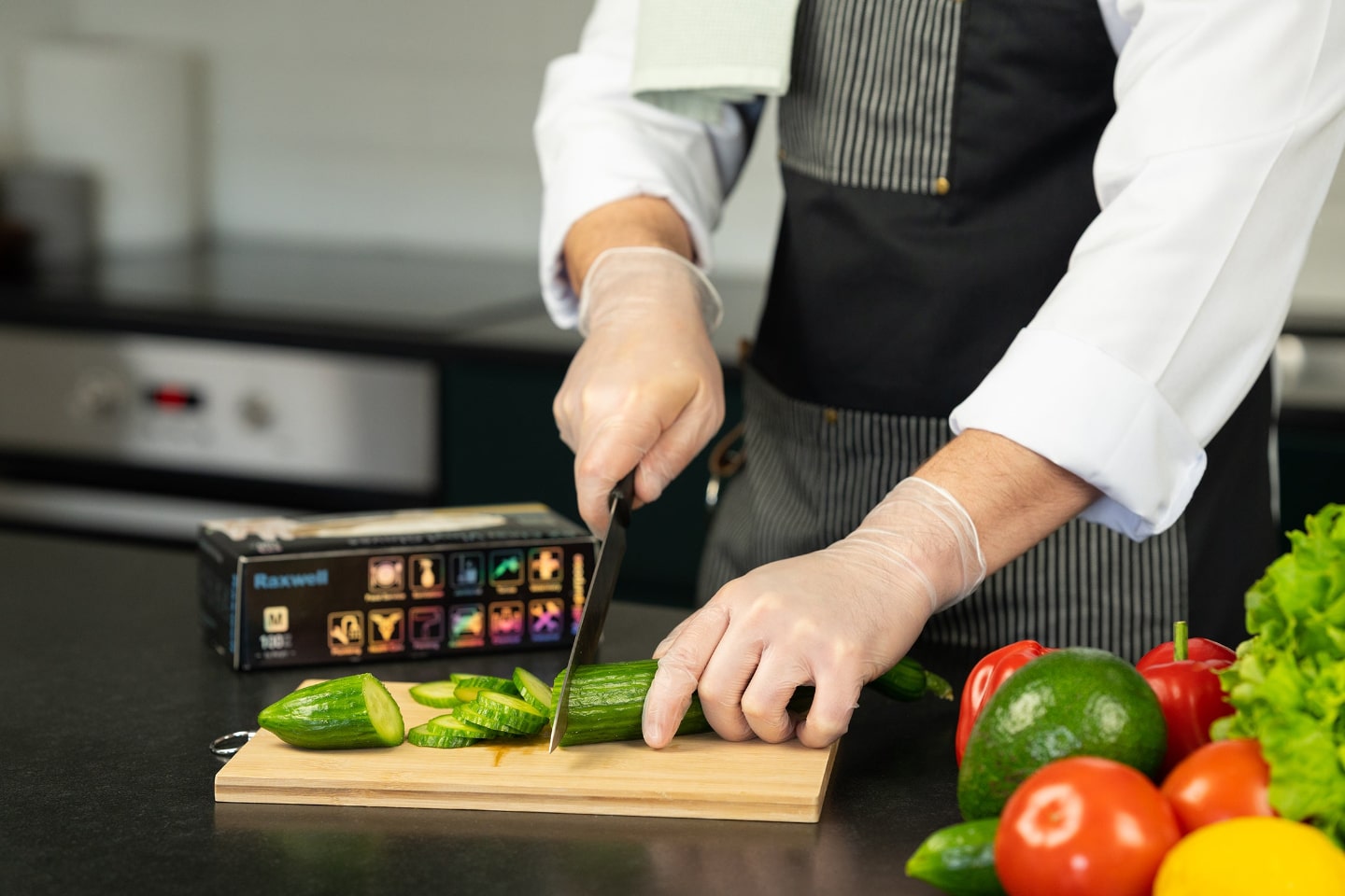 Professional cook slices cucumbers while wearing clear Raxwell vinyl gloves; a colorful Raxwell glove box sits nearby with fresh produce. Image conveys hygienic food prep.