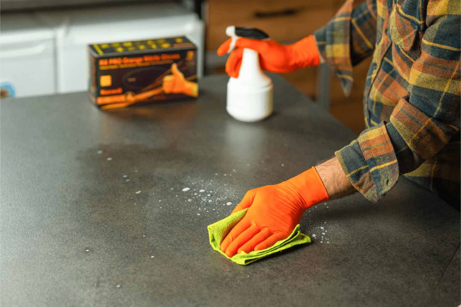 Person in orange Raxwell diamond-textured gloves sprays cleaner and wipes a countertop. The shot showcases non-slip grip and chemical safety.