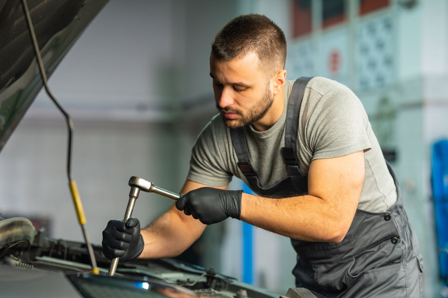 A male mechanic wearing gray overalls and black gloves uses a ratchet tool to work on a car engine in a garage or workshop. The car hood is open and the background is slightly blurred.