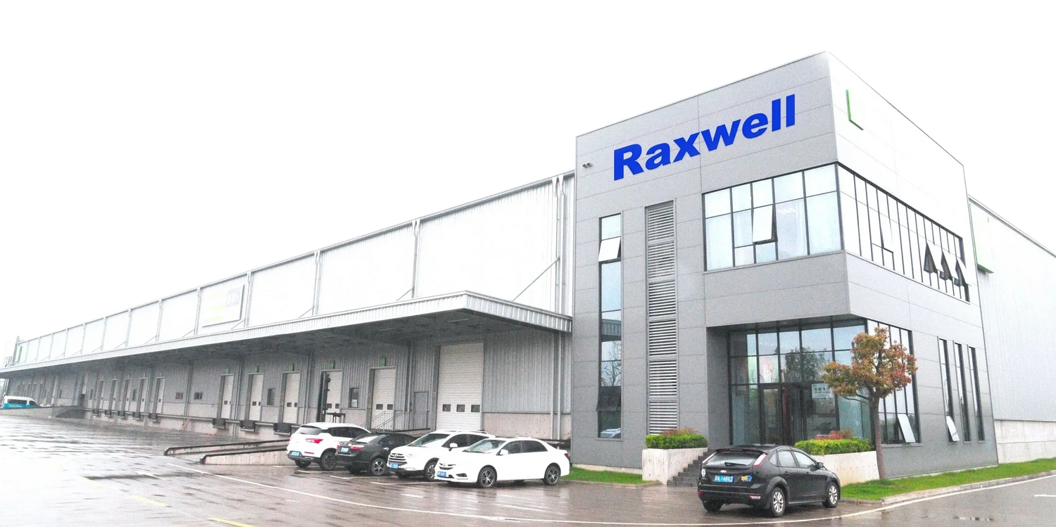 A modern industrial warehouse with large windows and the name Raxwell displayed on the building. Several cars are parked in front on a wet pavement.