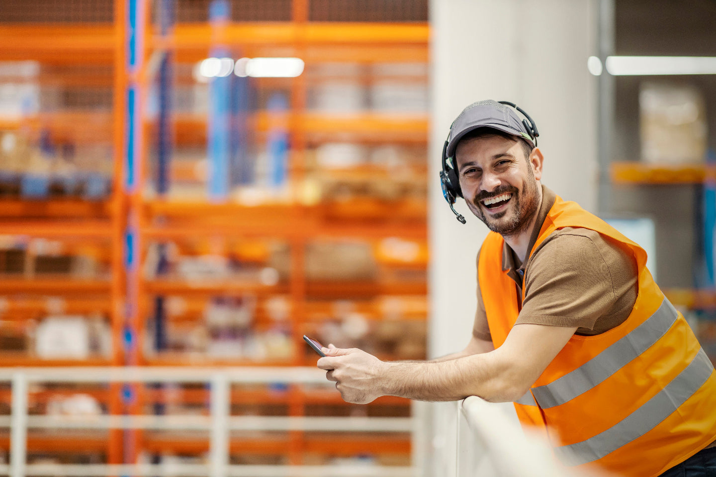 Smiling warehouse associate in orange hi-vis vest and headset leans on railing while holding a phone. Used by Raxwell as a friendly “contact us” visual.