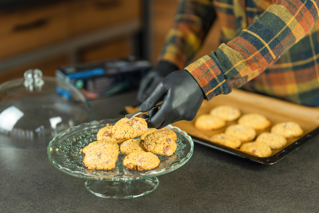 Baker plates cookies with tongs while wearing black Raxwell nitrile gloves. Scene demonstrates food-safe grip in home kitchens.