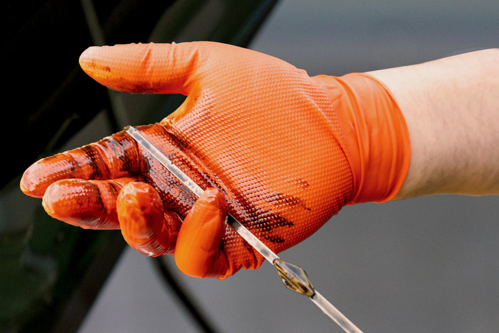 Close-up of an orange Raxwell glove coated in motor oil while checking a dipstick. Macro view emphasizes texture and barrier strength.