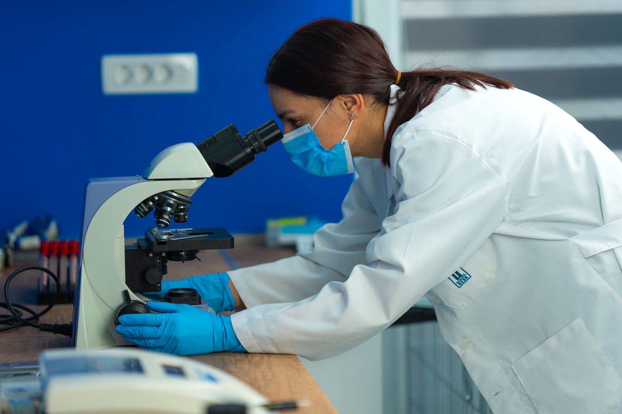 Scientist peers through a microscope wearing blue Raxwell gloves. Image underlines tactile sensitivity for precise lab work.