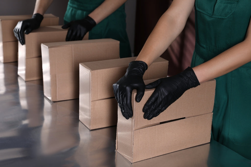 Workers seal cardboard boxes on an assembly line using black Raxwell gloves. Photo underlines secure grip for warehouse packing.