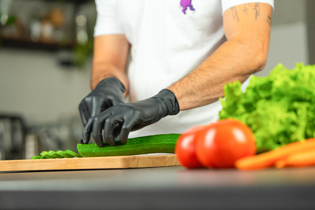 Home cook slices cucumber on a wooden board while wearing black Raxwell gloves. Fresh veggies reinforce safe food prep.