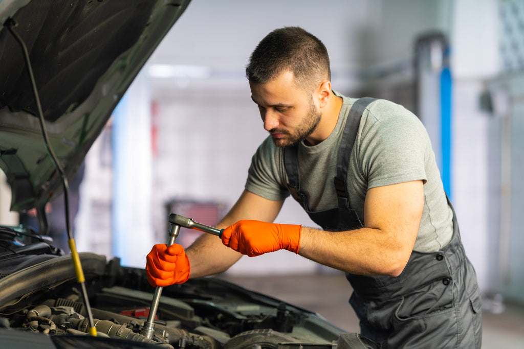 Mechanic uses a torque wrench wearing durable orange Raxwell gloves. Scene highlights high-visibility comfort for long repairs.