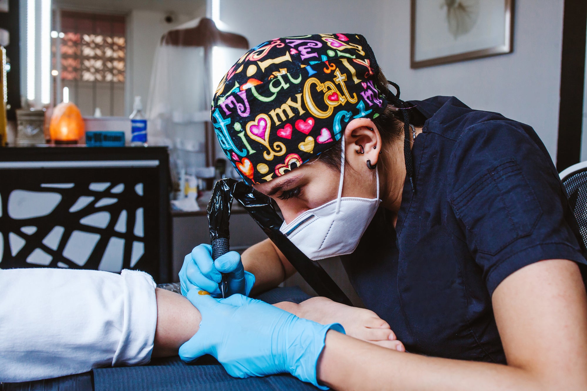Tattooist applies forearm art while wearing blue Raxwell gloves and a colorful scrub cap. Scene showcases hygienic barriers in creative studios.