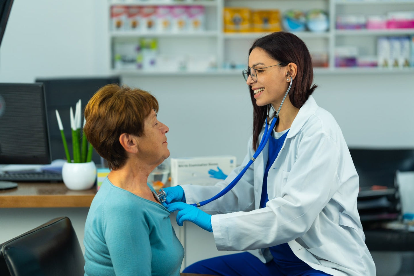 A doctor wearing a white coat and gloves uses a stethoscope to listen to an older womans chest in a medical office. Both are smiling, and shelves with medical supplies are visible in the background.