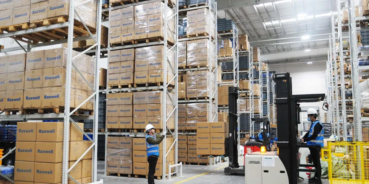 Workers in safety vests and helmets operate forklifts and walk between tall racks stacked with pallets of boxed goods in a brightly lit warehouse.