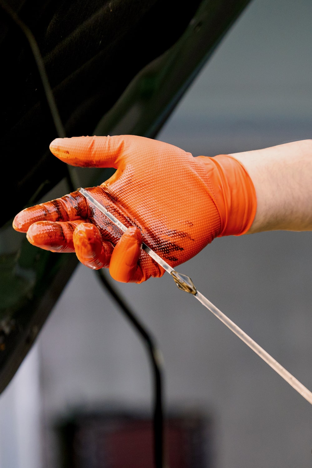 Mechanic’s hand in textured orange Raxwell glove holds an oil-coated dipstick under a car hood. Shows gloves standing up to greasy automotive work.