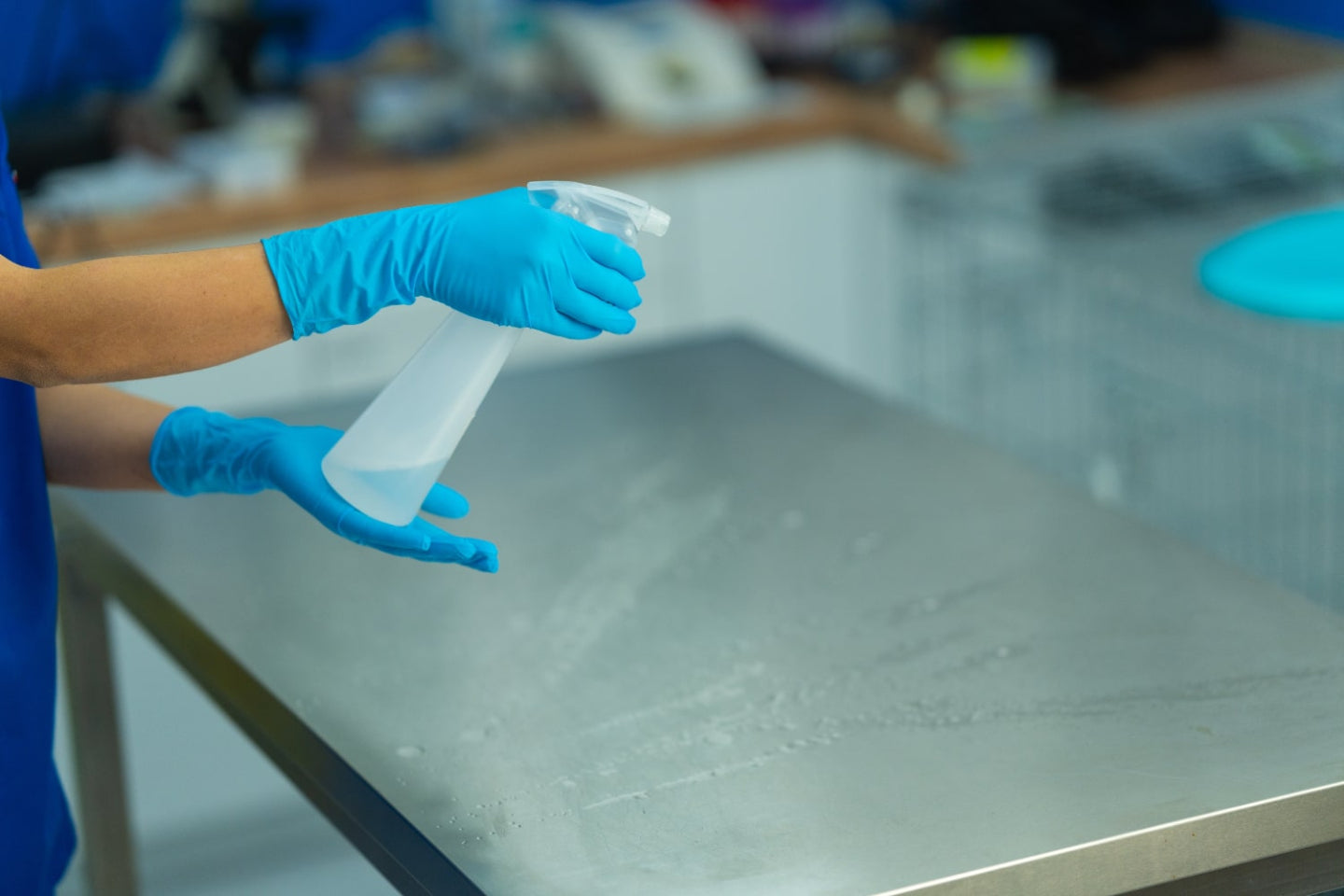 A person wearing blue gloves sprays disinfectant from a clear spray bottle onto a metal table in a clean, bright room, likely preparing the surface for sanitation.